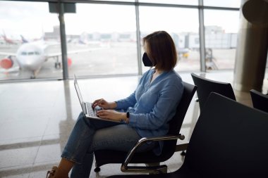Young woman freelancer wearing face mask working with a laptop lounge of airport while is waiting a departure of flight. Freelance business