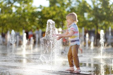 Küçük çocuk, güneşli yaz gününde kuru çeşmedeki su jetlerinin arasında oynuyor. Şehirdeki çocuklar için aktif yaz tatili.