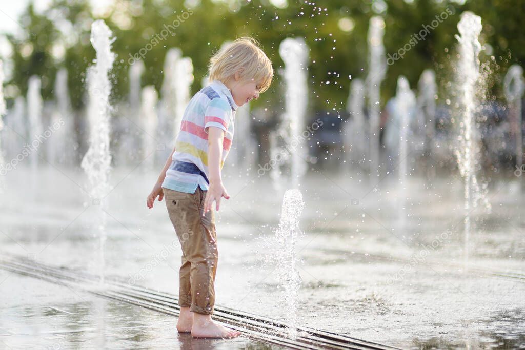 Little boy plays in the square between the water jets in the dry fountain at sunny summer day. Active summer leisure for kids in the city.