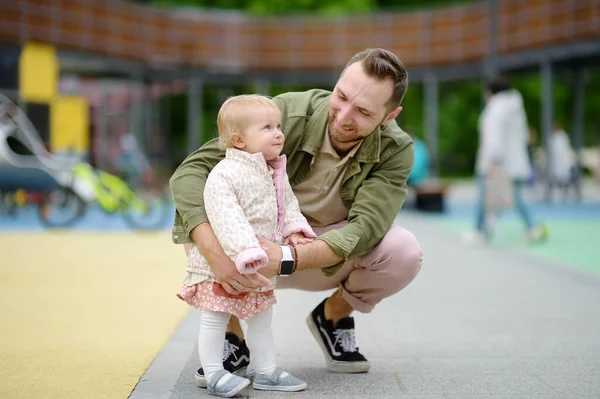 屋外の遊び場で楽しんでいるかわいい幼児の女の子 幼い父親は娘に歩くように教えている 赤ちゃんの第一歩 小さな子供を持つ家族のために歩く 幸せな親 — ストック写真
