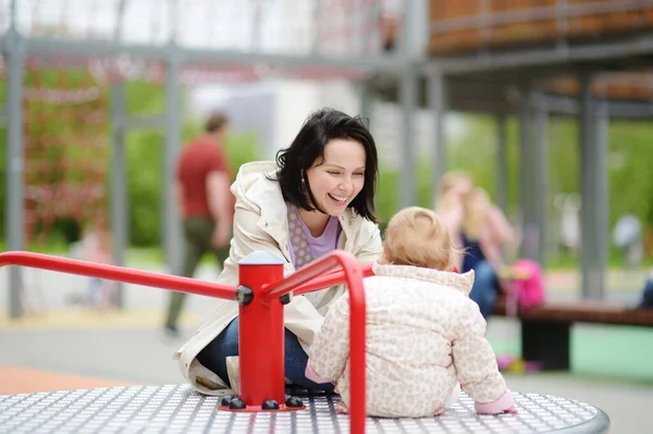 屋外の遊び場で楽しんでいる幼児の女の子 幸せな若い母親はカルーセルに赤ちゃんに乗る 秋小さな子供を持つ家族のためのアクティブなレジャー 幸せな母親 — ストック写真