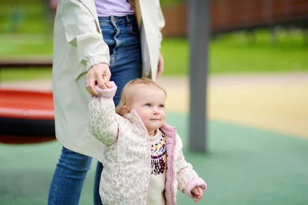 屋外の遊び場で楽しんでいるかわいい幼児の女の子 若い母親は娘に歩くように教えている 赤ちゃんの第一歩 小さな子供を持つ家族のために歩く 幸せな母親 — ストック写真