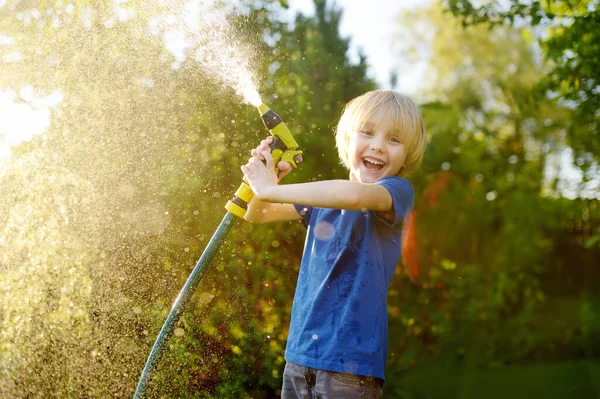 面白い小さな男の子は植物を散水し 日当たりの良い裏庭でスプリンクラーで庭のホースで遊んでいます 水のスプレーで楽しんでいる未就学児 子供のための夏の屋外活動 幸せな子供時代 — ストック写真