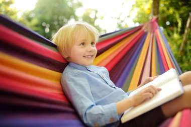 Küçük sevimli sarışın beyaz çocuk kitap okuyor ve arka bahçede ya da dışarıda renkli hamakla eğleniyor. Çocuklar için yaz tatili. Hamakta sallanan ve rahatlatan çocuk.