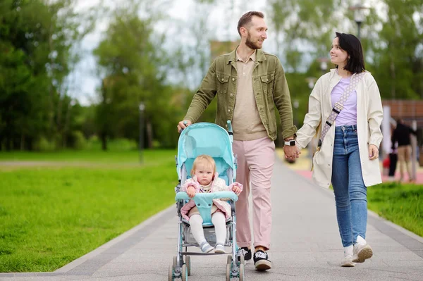 可爱的蹒跚学步的小女孩和她年轻可爱的父母散步时 骑着婴儿车 为有小孩的家庭提供积极的闲暇和散步 快乐的父母 — 图库照片