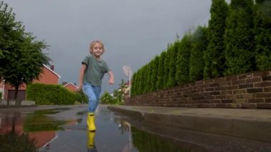 Küçük bir kasabada yağmurlu bir yaz gününde sarı lastik çizme giymiş, su birikintisine atlayan küçük bir çocuğun yavaş çekim videosu. Çocuk eğleniyor. Yağmurda çocuklar için açık hava oyunları.