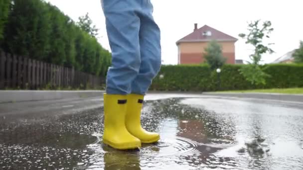 小さな町で雨の夏の日に水の水たまりにジャンプ黄色のゴムブーツを着て小さな男の子のスローモーションビデオ 子供は楽しんでる 雨の中で子供のための屋外ゲーム — ストック動画
