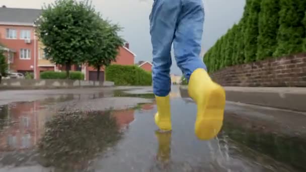 在小镇的下雨天 穿着黄色橡胶靴的小男孩在水坑里跳跃的慢镜头 孩子们玩得很开心雨天儿童户外运动 — 图库视频影像