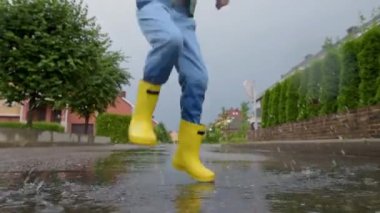 Küçük çocuk, yağmurlu bir yaz gününde küçük bir kasabada su birikintisine atlayan sarı lastik çizmeler giyiyor. Çocuk eğleniyor. Yağmurda çocuklar için açık hava oyunları.