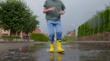 Küçük bir kasabada yağmurlu bir yaz gününde sarı lastik çizme giyen ve su birikintisine atlayan küçük bir çocuğun yavaş çekim videosu. Çocuk eğleniyor. Yağmurda çocuklar için açık hava oyunları.