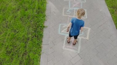 Asfalta çizilmiş seksek eşliğinde zıplayan küçük bir çocuğun hava manzarası. Çocuk bahar günü oyun bahçesinde seksek oynuyor. Üst Manzara. Çocuklar için açık hava aktiviteleri.