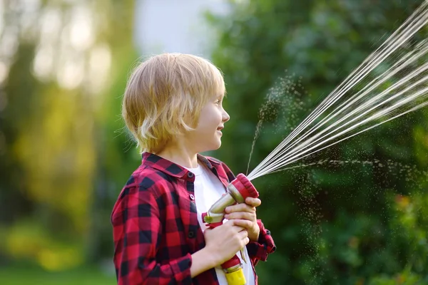 Funny little boy watering plants and playing with garden hose with sprinkler in sunny backyard. Preschooler child having fun with spray of water. Summer outdoors activity for kids.