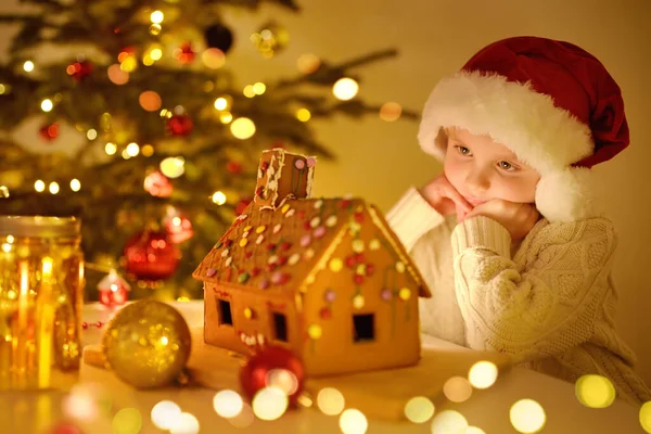 小男孩欣赏着甜蜜的自制姜饼屋装饰着糖果和釉面 姜饼屋矗立在圣诞树旁的桌子上 桌上点缀着装饰灯 蜡烛和节日的花环 — 图库照片