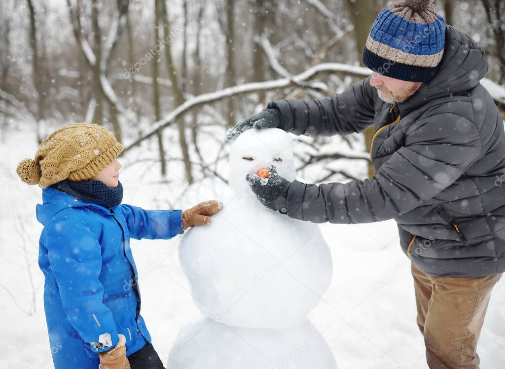 Little boy with his father building snowman in snowy park. Active outdoors leisure with family with children in winter. Kid during stroll in a snowy winter park