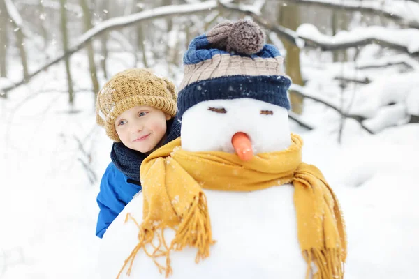 小男孩在雪地公园里堆雪人 孩子们抱着戴着帽子和围巾的雪人 冬天与有孩子的家庭一起积极地进行户外活动 孩子们在雪地的冬日公园里散步 — 图库照片