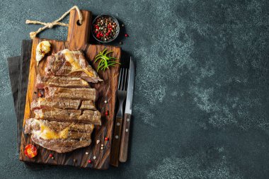 Closeup ready to eat steak ribeye breeds of black Angus with herbs, garlic and butter on a wooden Board. The finished dish for dinner on a dark stone background. Top view with copy space