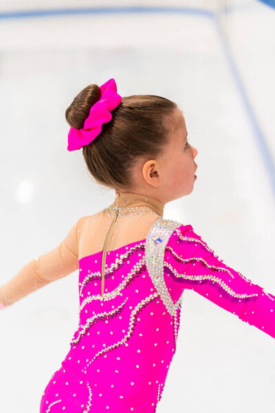 Маленькая фигуристка в розовом платье практикуется на крытом катке.