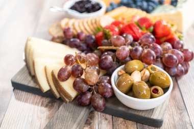 Aperatif olarak sunulan tahtanın üzerinde gurme peyniri, kraker ve meyve var..