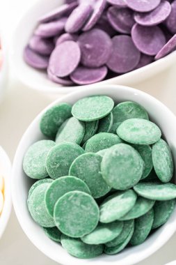 Beyaz arka planda çikolata şekillerini eritmek için beyaz kaselerde yuvarlak çikolata parçacıklarının gökkuşağı renkleri.