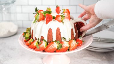 Step by step. Slicing red velvet bundt cake with cream cheese frosting garnished with fresh strawberries.