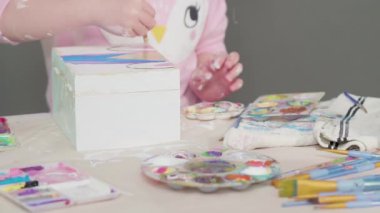 Küçük kız ahşap bir kutuya akrilik boyalı beyaz bir tek boynuzlu at çiziyor..