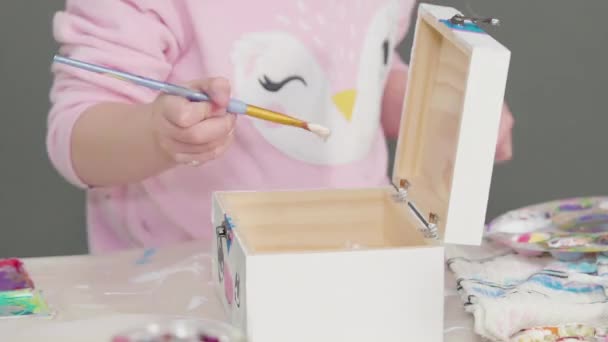 小さな女の子は木製の箱の上にアクリル絵具で白いユニコーンを描く — ストック動画