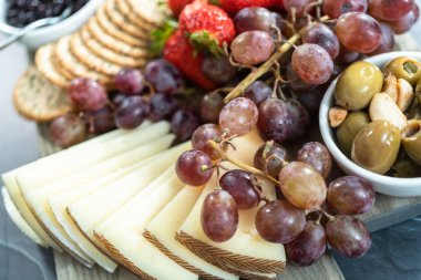 Aperatif olarak sunulan tahtanın üzerinde gurme peyniri, kraker ve meyve var..