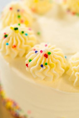 Üzerinde gökkuşağı parçacıklarıyla süslenmiş yedi tane şekerleme olan orta boy bir pasta üst kenarı. Altın pasta tahtası kısmen görülebilir. Işık ayarı canlı ve şenlikli bir hava yaratıyor..