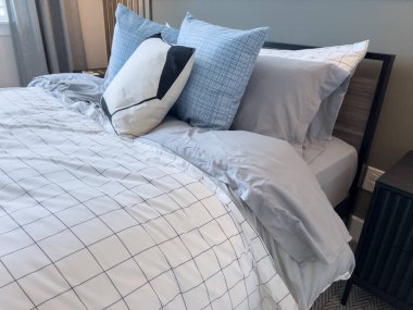 Yatak odasındaki kareli yatak odası, nötr gri yastıklar ve yastıklı yatak başlığı. Doğal ışık pencereden girer..