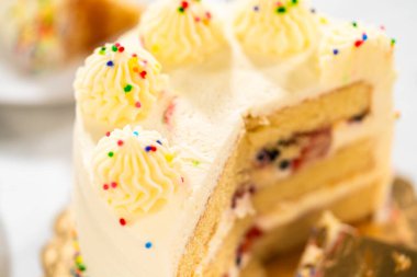 A cake with a missing slice sits on a golden board, showing its inner fruit-filled layers and piped frosting. The top is decorated with sprinkles and soft peaks of icing.