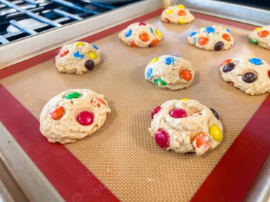 Çikolata şekerli renkli kurabiye hamuru topları pişirme tepsisinin içindeki silikon bir paspasın üzerinde düzenlenir. Fırın ışığı onların canlı görüntülerini vurgular..