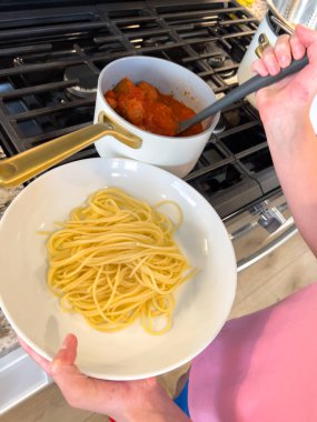 Pembe gömlekli bir kişi tencereden spagettiyi sol elinde tutarken beyaz bir tabağa koyar. Köfteli fırın ve tencere arka planda gün ışığında görülebiliyor.