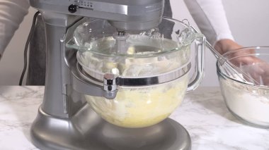 Bir karıştırıcı mermer tezgahın üzerine yerleştirilmiş cam bir kasede kremalı kurabiye hamuru olarak gösterilir. Malzemeler temiz bir mutfak ortamında parlak ışık altında harmanlanıyor..