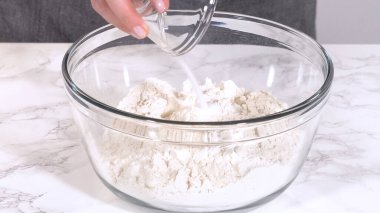 Tuz, mermer tezgahtaki un dolu cam bir kaseye dökülüyor. Sahne, kuru malzemelerin parlak ışık altında birleştirilmesinin basamaklarını yakalıyor..