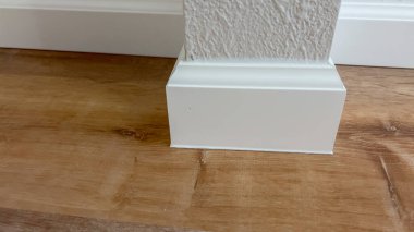 Orta tonda ahşap döşemeye karşı beyaz süpürgelik köşesi detaylarının yakın plan görüntüsü. Işık yumuşak ve doğal, duvarla zemin arasındaki keskin mimari kontrastı vurguluyor..