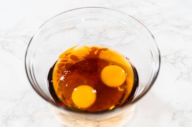 İki çiğ yumurta ve akçaağaç pekmezi içeren cam kasenin üst görüntüsü Amber Zencefilli Kurabiye Hamuru için koyu bir temel karışımı oluşturuyor..