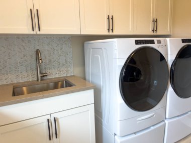 İçinde küçük bir lavabo, çamaşır makinesi ve beyaz dolaplar olan modern çamaşır odası. Gündüz, çağdaş bir iç mekanda temiz çizgiler ve basitliği vurgular..