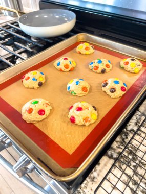 Çikolata şekerli renkli pişmemiş kurabiyeler silikon kaplı bir pişirme tepsisi üzerinde düzenlenir. Tepsi bir fırının içine konur ve kapı açılır. Parlak mutfak ışığı altında pişirilmeye hazırdır..