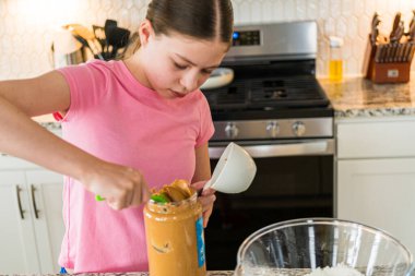 Pembe tişörtlü bir kız büyük bir kavanozdan fıstık ezmesini dikkatlice ölçü kabına sokuyor. Mutfak, beyaz dolaplar ve granit tezgahlarla aydınlık ve modern..