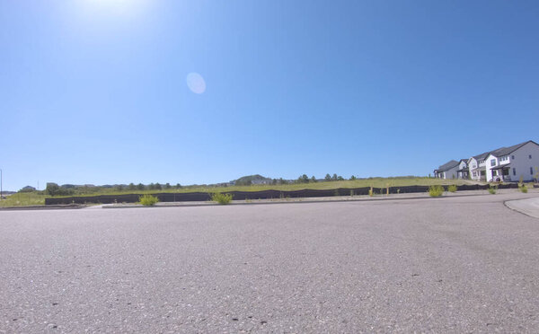 Открытый вид на окрестную улицу, граничащую с жилыми домами и пустыми полями. Обстановка кажется просторной и свежеразвитой, с ярким небом и широкими дорогами.