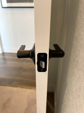 Bitişik odanın bir kısmını gösteren beyaz bir kapının siyah mandalının yakın çekim görüntüsü. Mat tamamlama donanımı temiz bir görünüm için boyalı yüzey ile zıt.