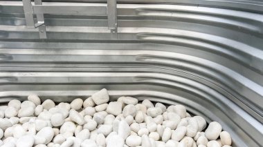 Bükülmüş metalden yapılmış ve küçük beyaz peyzaj taşlarıyla dolu bodrum penceresi. Görüntü doğal ışık altında çekiliyor, temiz ve asgari estetiği vurguluyor..