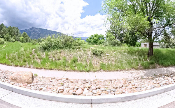 A sloping grassy area with decorative rocks along the sidewalk and curb. The background features trees and a wide cloudy sky, creating a natural suburban landscape.