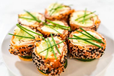 Beyaz bir tabakta ustaca hazırlanmış füme somon soslu krem peynirli rulo. Ekmekler canlı tütsülenmiş somona sarılmış, siyah ve beyaz susam tohumları serpiştirilmiş, ve en üstte