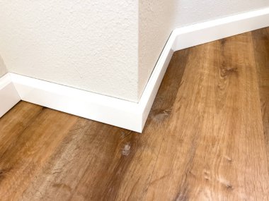 Gerçekçi ahşap döşeme üzerinde keskin açılı bir duvar köşesi takip eden beyaz bir süpürgeliğin ayrıntılı görüntüsü. Tasarım günlükleri veya döşeme materyali rehberleri için kullanışlı.