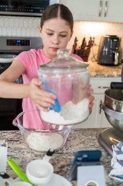 Genç kız, modern bir mutfakta malzemeleri pişirirken un dolu büyük bir cam kavanozu kaldırır. Fırın aletleri ve telefon standı tezgahın üzerinde..
