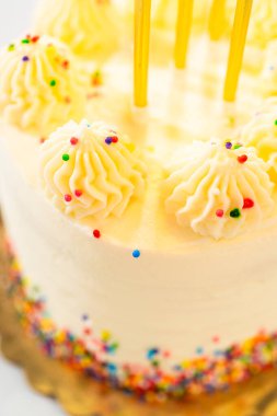 Yuvarlak bir doğum günü pastasını gösteren dikey bir görüntü. Boru şeklinde girdaplar ve uzun sarı mumlar. Pasta altın bir zeminde, altta çok renkli süslemelerle süslenmiş..