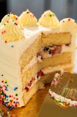 Boru şeklinde girdap ve gökkuşağı parçalı uzun vanilyalı pasta kısmen dilimlenmiş duruyor. Altın renkli bir spatula dilimin içinde duruyor. Arkaplan karşıtlık için bulanık koyu tonları gösterir.