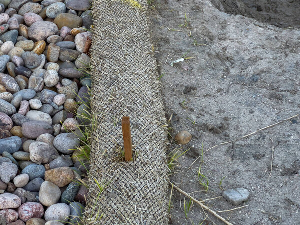 A closeup view shows a natural fiber erosion control mat held in place with metal stakes at the edge of a dirt area. Decorative rocks are visible beside the mat. The image captures soil stabilization