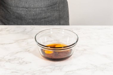 Amber zencefilli kurabiye hamurunun bir parçası olarak mermer tezgahın üzerine yerleştirilmiş yumurta ve akçaağaç pekmezli cam kasenin yan görünüşü, ıslak malzemelerin kombinasyonunu vurguluyor.